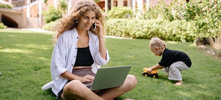 A woman talking over the phone with movers about moving from San Francisco to Portland while using a laptop and while a child is playing next to her